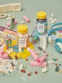 A colorful arrangement of nostalgic items including two jars labeled 'not pot' with gummies inside. Surrounding the jars are a Rubik's cube, hair rollers, scattered popcorn, a retro telephone handset, a comb, nail polish, headphones, various candies, and vintage-style cartoon cards. The background is a muted teal color.