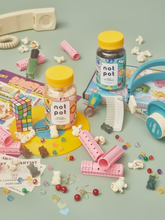 A colorful arrangement of nostalgic items including two jars labeled 'not pot' with gummies inside. Surrounding the jars are a Rubik's cube, hair rollers, scattered popcorn, a retro telephone handset, a comb, nail polish, headphones, various candies, and vintage-style cartoon cards. The background is a muted teal color.