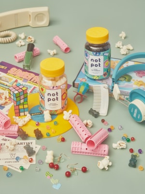 A colorful arrangement of nostalgic items including two jars labeled 'not pot' with gummies inside. Surrounding the jars are a Rubik's cube, hair rollers, scattered popcorn, a retro telephone handset, a comb, nail polish, headphones, various candies, and vintage-style cartoon cards. The background is a muted teal color.