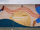 A colorful mural on a brick wall featuring abstract shapes and vibrant colors with text reading 'LOOKING PRETTY, MUSIC CITY.' It has flowing orange lines, a large yellow circle, and a combination of dark blue and brown elements. A no parking sign stands in front of it, casting a shadow on the sidewalk.