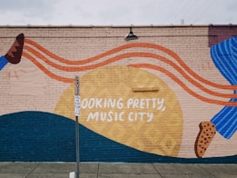 A colorful mural on a brick wall featuring abstract shapes and vibrant colors with text reading 'LOOKING PRETTY, MUSIC CITY.' It has flowing orange lines, a large yellow circle, and a combination of dark blue and brown elements. A no parking sign stands in front of it, casting a shadow on the sidewalk.