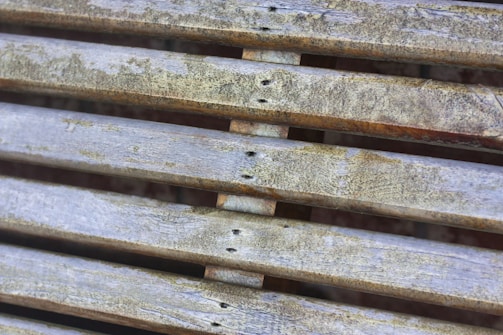 Weathered wooden slats are arranged horizontally, displaying signs of aging with faded paint and patches of moss or dirt. The surface texture is rough and uneven, indicating exposure to the elements over time.
