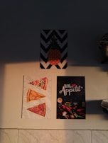 Three posters are arranged on a wall. The top poster features a pineapple on a black and white zigzag background. The bottom left poster displays three slices of pizza with different toppings, positioned on a white textured background. The bottom right poster reads 'Bon Appetit' in decorative lettering and features various vegetables and pasta scattered on a dark background.