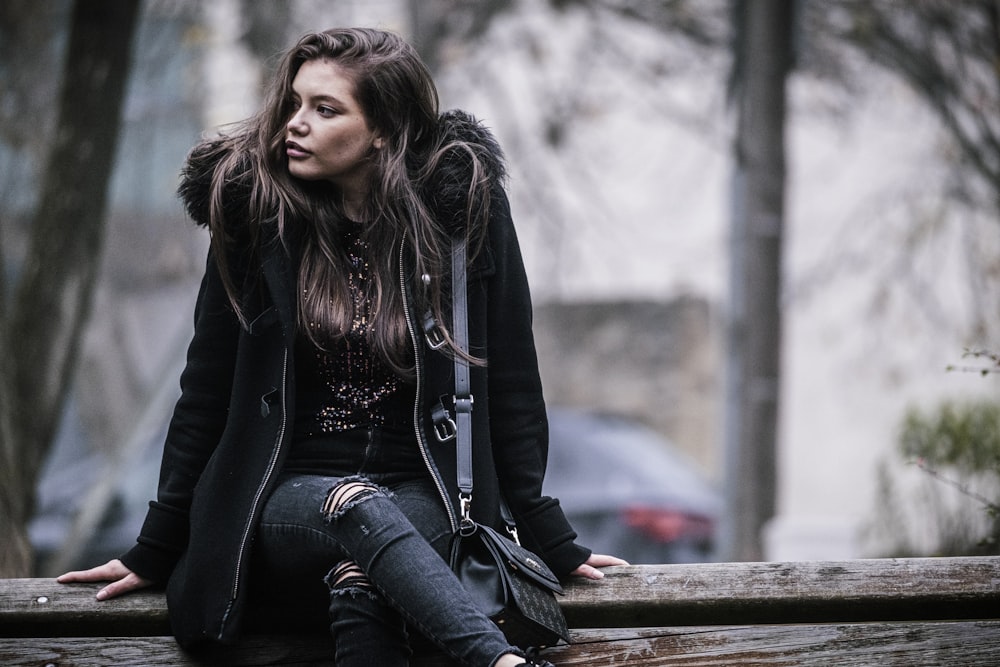 woman in black jacket and blue denim jeans sitting on brown wooden bench during daytime