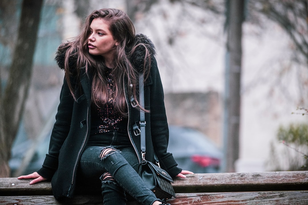 woman in black jacket and blue denim jeans sitting on brown wooden bench during daytime