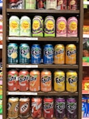 A variety of soda cans are arranged on shelves. The top shelf features energy drinks and Sparkling Chupa Chups in various flavors. The second and third shelves display rows of Fanta cans in different flavors such as lime, berry, fruit punch, peach, and pineapple. The bottom shelf contains A&W Cream Soda, Cherry 7 Up, and additional flavors of Fanta.