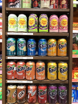 A variety of soda cans are arranged on shelves. The top shelf features energy drinks and Sparkling Chupa Chups in various flavors. The second and third shelves display rows of Fanta cans in different flavors such as lime, berry, fruit punch, peach, and pineapple. The bottom shelf contains A&W Cream Soda, Cherry 7 Up, and additional flavors of Fanta.