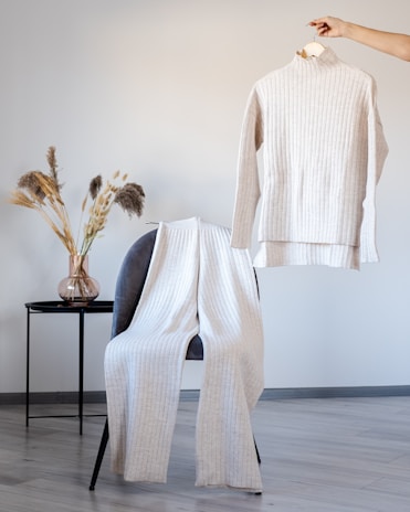 A beige ribbed sweater and pants set are displayed in a minimalist setting. The sweater is being held on a hanger by a hand, while the pants are draped over a grey chair. Next to the chair, dried decorative grasses are arranged in a transparent vase on a small black table. The floor is light wood, and the wall is a soft grey color.