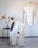 A beige ribbed sweater and pants set are displayed in a minimalist setting. The sweater is being held on a hanger by a hand, while the pants are draped over a grey chair. Next to the chair, dried decorative grasses are arranged in a transparent vase on a small black table. The floor is light wood, and the wall is a soft grey color.