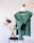 A green knitted sweater is held up on a hanger by a hand. In the background, there is a minimalist setting with dried plants in a transparent vase placed on a black round table against a neutral wall.