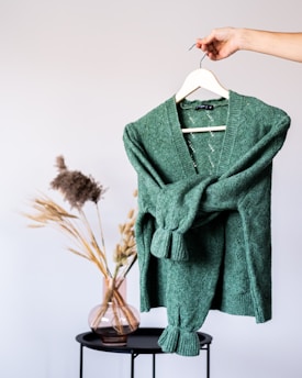 A green knitted sweater is held up on a hanger by a hand. In the background, there is a minimalist setting with dried plants in a transparent vase placed on a black round table against a neutral wall.