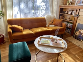 A cozy living room with a mustard-colored sofa adorned with pillows and a furry blanket. A small side table nearby has a tray with decorative items, including a bowl with smoldering sage. The room features wooden shelves filled with books and decorative pieces, and artwork leaning against a tiled fireplace. Sunlight filters through sheer curtains, casting shadows on the wooden floor.