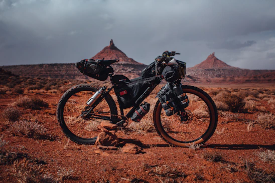A rugged mountain bike resting against volcanic rocks under a cloudy sky, maps and GPS gear scattered nearby.
