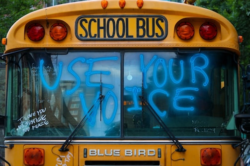 A school bus with the words 'USE YOUR VOICE' written in bright blue marker on the front windshield. Other smaller messages like 'I LOVE YOU', 'NO JUSTICE', and 'NO PEACE' are also visible. The bus has a yellow exterior with flashing orange lights and the label 'SCHOOL BUS' above the windshield.