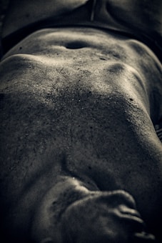 A close-up, moody black-and-white photograph highlighting the muscular torso and abdomen of a person. The skin texture and muscle definition are accentuated by dramatic lighting and shadows.