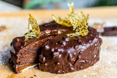 A rich, chocolate-covered dessert with a smooth, glossy texture on its exterior. The cake is topped with intricate golden sugar decorations and is sliced to reveal a dense, fudgy interior. It rests on a wooden surface dusted with a light layer of powdered sugar.