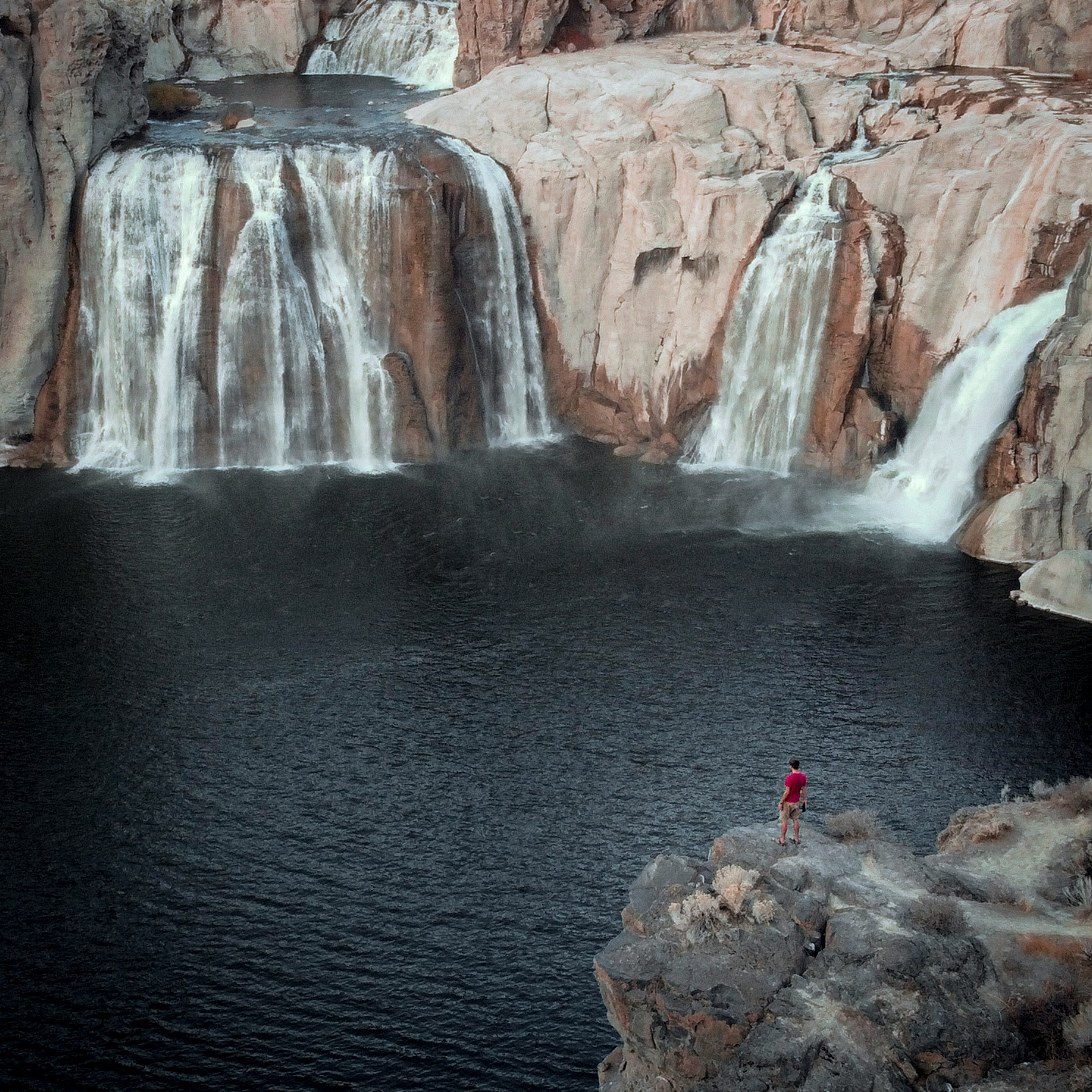 person in red jacket standing on rock near waterfalls during daytime