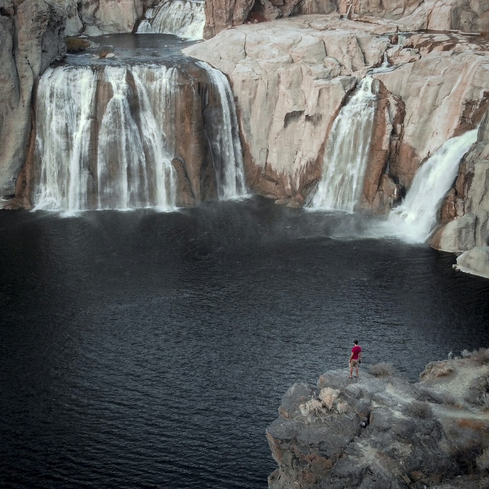 person in red jacket standing on rock near waterfalls during daytime