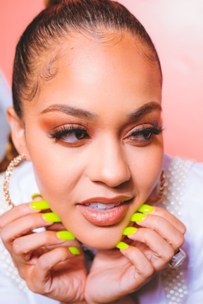 A close-up portrait of a person with neatly styled hair pulled back, wearing bright neon green nail polish. The individual has detailed makeup with prominent false eyelashes and is adorned with pearl jewelry.