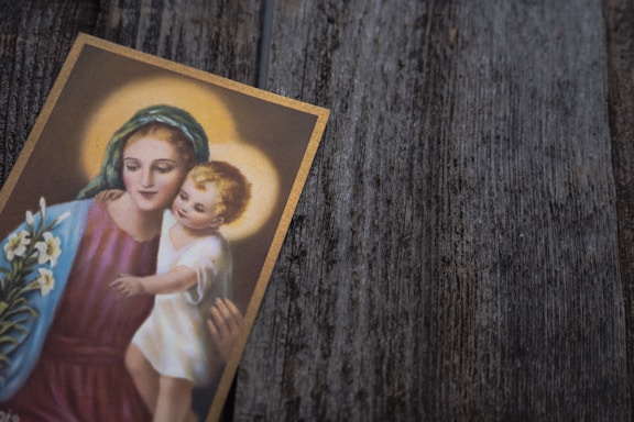A card featuring an artistic depiction of a mother and child with halos, reflecting a serene and calm expression. The mother wears a blue-green headscarf and a purple garment, while holding the child in her arms. A bouquet of white lilies is also visible at the side.