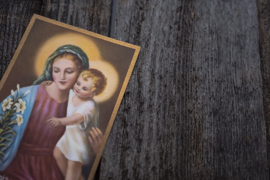 A card featuring an artistic depiction of a mother and child with halos, reflecting a serene and calm expression. The mother wears a blue-green headscarf and a purple garment, while holding the child in her arms. A bouquet of white lilies is also visible at the side.