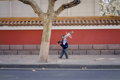A person walks down a sidewalk carrying a bundle of cleaning tools over their shoulder. The background features a traditional-style wall painted red with ornate tile roofing, adding a contrasting charm to the urban setting. Scattered leaves are visible on the pavement and street, indicating an autumn season.