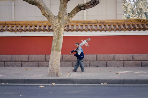 A person walks down a sidewalk carrying a bundle of cleaning tools over their shoulder. The background features a traditional-style wall painted red with ornate tile roofing, adding a contrasting charm to the urban setting. Scattered leaves are visible on the pavement and street, indicating an autumn season.