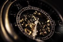 A close-up view of the intricate inner workings of a mechanical watch. The prominent focus is on the golden gears and the hands of the watch, which are finely detailed. The lighting highlights the metallic surfaces, creating a reflective and luxurious appearance.