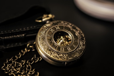 An ornate brass pocket watch featuring intricate engravings and Roman numerals around its face. The watch is attached to a chain and placed on a dark surface, adding to its vintage aesthetic.