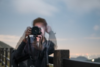 A blurred image of a person holding a camera against a backdrop of a railing and a scenic horizon. The motion blur suggests movement, creating a ghostly effect.