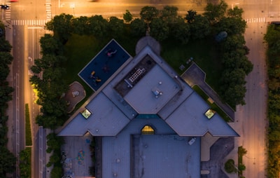 An aerial view of a large building with a complex, geometric rooftop design surrounded by greenery and streets. A playground with equipment is visible next to the building, and there are well-lit roads with moving vehicles on two sides. Trees form a border around the property, and there are crosswalks and sidewalks on the streets.