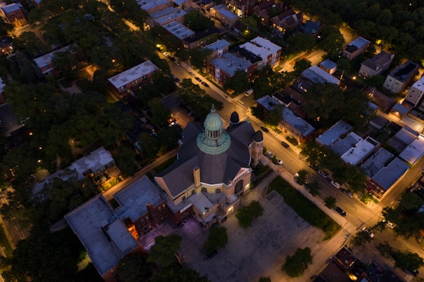 An aerial view of an illuminated urban area featuring a prominent church with a green dome surrounded by residential buildings and lined streets. Trees and streetlights enhance the nighttime cityscape, casting a warm glow over the neighborhood.