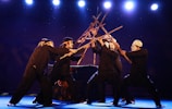 A group of performers dressed in black attire are engaged in a theatrical performance using wooden sticks, creating an intricate formation. They wear white masks and headbands, standing on a stage illuminated by bright spotlights against a dark background.