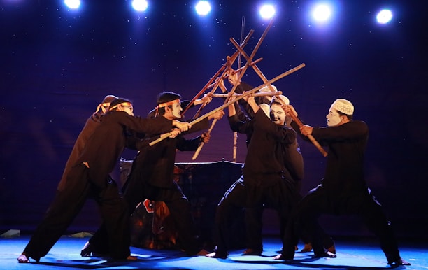 A group of performers dressed in black attire are engaged in a theatrical performance using wooden sticks, creating an intricate formation. They wear white masks and headbands, standing on a stage illuminated by bright spotlights against a dark background.