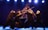 A group of performers dressed in black attire are engaged in a theatrical performance using wooden sticks, creating an intricate formation. They wear white masks and headbands, standing on a stage illuminated by bright spotlights against a dark background.