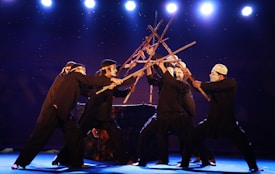 A group of performers dressed in black attire are engaged in a theatrical performance using wooden sticks, creating an intricate formation. They wear white masks and headbands, standing on a stage illuminated by bright spotlights against a dark background.