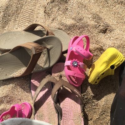 A collection of various sandals and flip-flops resting on sandy ground. The footwear includes a pair of brown flip-flops, a pair of pink sandals decorated with a floral detail, a smaller pink sandal, and a bright yellow clog-type shoe.