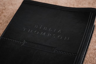 A close-up view of a book cover, embossed with the words 'Bíblia Thompson.' The cover appears to be made of dark, textured leather, giving it a classic and refined look. It rests on a light brown surface that adds contrast to the dark cover.