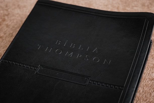 A close-up view of a book cover, embossed with the words 'Bíblia Thompson.' The cover appears to be made of dark, textured leather, giving it a classic and refined look. It rests on a light brown surface that adds contrast to the dark cover.