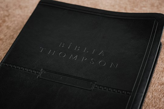 A close-up view of a book cover, embossed with the words 'B&iacute;blia Thompson.' The cover appears to be made of dark, textured leather, giving it a classic and refined look. It rests on a light brown surface that adds contrast to the dark cover.