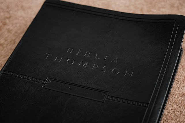 A close-up view of a book cover, embossed with the words 'B&iacute;blia Thompson.' The cover appears to be made of dark, textured leather, giving it a classic and refined look. It rests on a light brown surface that adds contrast to the dark cover.