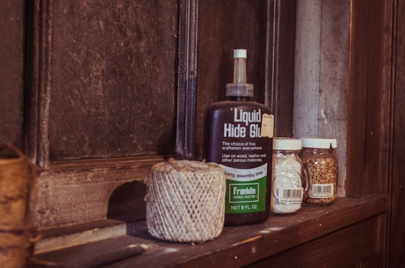 A wooden shelf holds a bottle of 'Liquid Hide Glue', a roll of twine, and two jars. One jar appears to contain small grains or seeds, and the other holds a white substance. The setting suggests a rustic or vintage atmosphere.