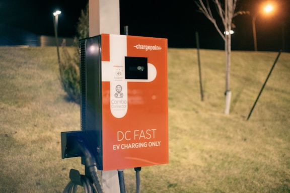 An electric vehicle charging station featuring a red and white design. The station is labeled with 'DC Fast EV Charging Only' and includes a combo connector. It is situated outdoors on a grassy area during nighttime, illuminated by nearby artificial lights.