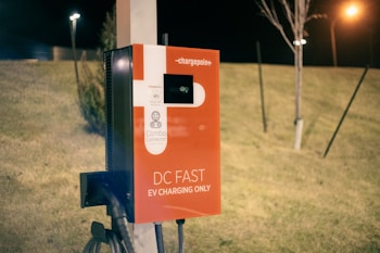 An electric vehicle charging station featuring a red and white design. The station is labeled with 'DC Fast EV Charging Only' and includes a combo connector. It is situated outdoors on a grassy area during nighttime, illuminated by nearby artificial lights.
