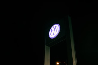 A tall sign displaying the Volkswagen logo is illuminated against a dark night sky. The sign is predominantly blue and white, providing a stark contrast against the black background.