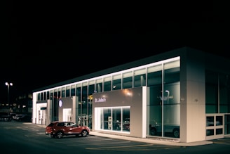 A car dealership at night with bright lights illuminating the building's modern, glass-front design. A red SUV is parked near the entrance, and the interior showcases more vehicles. The dealership is branded with the Volkswagen logo and the name St. John's is visible.