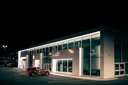 A car dealership at night with bright lights illuminating the building's modern, glass-front design. A red SUV is parked near the entrance, and the interior showcases more vehicles. The dealership is branded with the Volkswagen logo and the name St. John's is visible.