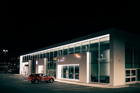 A car dealership at night with bright lights illuminating the building's modern, glass-front design. A red SUV is parked near the entrance, and the interior showcases more vehicles. The dealership is branded with the Volkswagen logo and the name St. John's is visible.