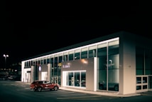 A car dealership at night with bright lights illuminating the building's modern, glass-front design. A red SUV is parked near the entrance, and the interior showcases more vehicles. The dealership is branded with the Volkswagen logo and the name St. John's is visible.