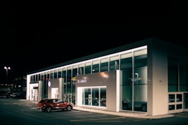 A car dealership at night with bright lights illuminating the building's modern, glass-front design. A red SUV is parked near the entrance, and the interior showcases more vehicles. The dealership is branded with the Volkswagen logo and the name St. John's is visible.
